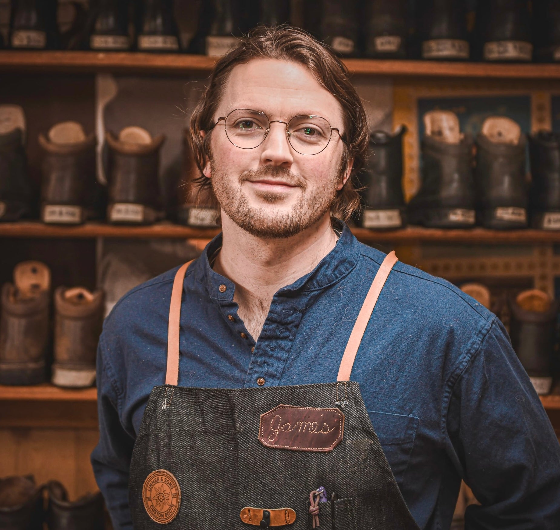Man wearing a denim apron with 'James' embroidered on it, standing in front of shelves with leather boots.